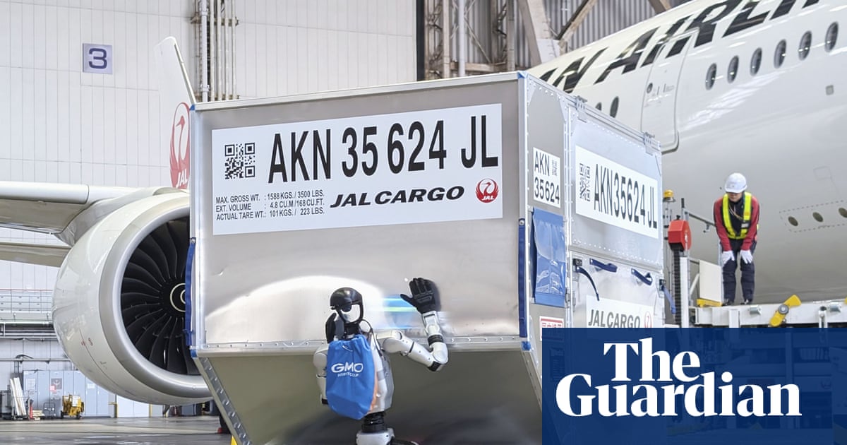 A humanoid robot tentatively pushing a piece of luggage onto a conveyor belt next to a Japan Airlines passenger plane.